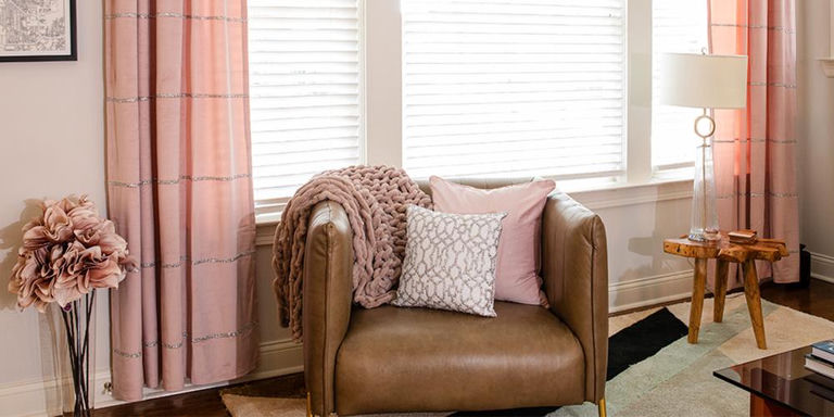 Leather armchair with blush pillows, knit throw, pink curtains, and a side lamp