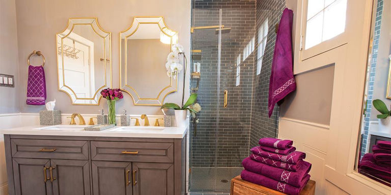 Bathroom vanity with gold mirrors and faucets, gray tile shower, and purple towels