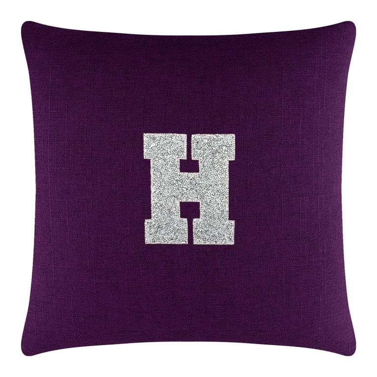 Purple square pillow featuring a rhinestone-embellished letter "H" in silver on a textured fabric surface.