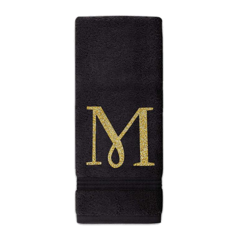 Black plush cotton terry hand towel with a glittering gold rhinestone monogram "M" and decorative trim at the bottom.