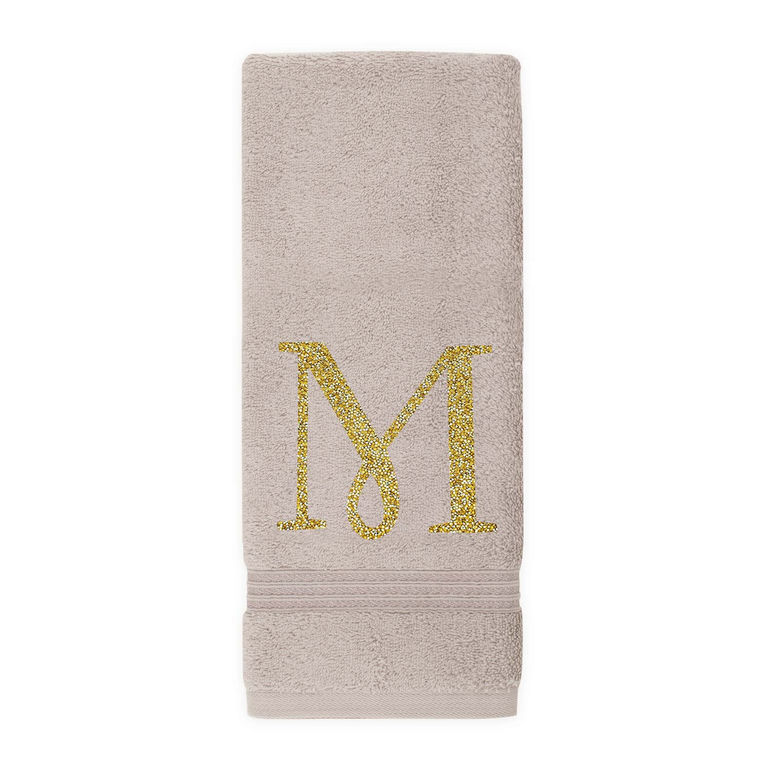 Taupe cotton terry hand towel with a gold rhinestone monogram "M" and decorative trim at the bottom.