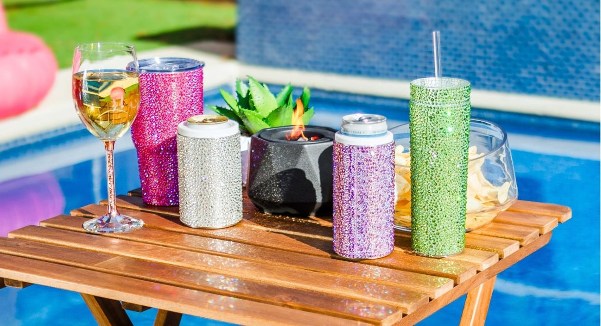 Colorful rhinestone-studded drinkware and can holders arranged on a wooden table by a pool, including a wine glass,  and sparkling tumblers