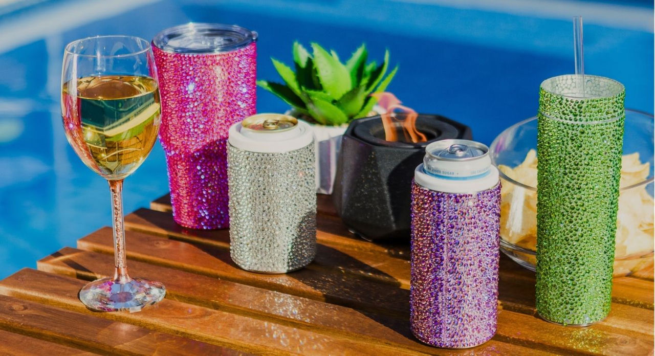 Colorful rhinestone-studded drinkware and can holders arranged on a wooden table by a pool, including a wine glass,  and sparkling tumblers