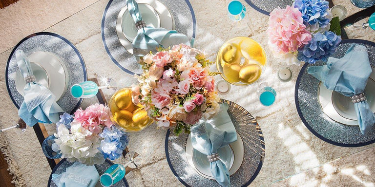 Elegante mesa de comedor temática de primavera con manteles plateados brillantes, vajilla blanca y servilletas azules claras adornadas con anillos de servilleta de pedrería. La mesa de espejo presenta centros florales pastel con hortensias rosas, blancas y azules, cuencos de vidrio llenos de limones amarillos y cócteles turquesas en vasos teñidos de azul. La suave alfombra crema debajo agrega un contraste acogedor a la decoración glamorosa de la mesa.