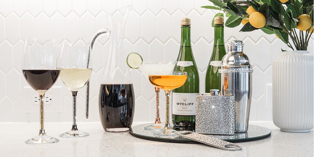 A&amp;&#x23;x20&#x3B;sophisticated&amp;&#x23;x20&#x3B;bar&amp;&#x23;x20&#x3B;cart&amp;&#x23;x20&#x3B;setup&amp;&#x23;x20&#x3B;featuring&amp;&#x23;x20&#x3B;an&amp;&#x23;x20&#x3B;array&amp;&#x23;x20&#x3B;of&amp;&#x23;x20&#x3B;cocktails&amp;&#x23;x20&#x3B;and&amp;&#x23;x20&#x3B;spirits.&amp;&#x23;x20&#x3B;Various&amp;&#x23;x20&#x3B;glasses&amp;&#x23;x20&#x3B;hold&amp;&#x23;x20&#x3B;different&amp;&#x23;x20&#x3B;colored&amp;&#x23;x20&#x3B;drinks&amp;&#x23;x20&#x3B;including&amp;&#x23;x20&#x3B;red&amp;&#x23;x20&#x3B;and&amp;&#x23;x20&#x3B;white&amp;&#x23;x20&#x3B;wine,&amp;&#x23;x20&#x3B;an&amp;&#x23;x20&#x3B;amber-colored&amp;&#x23;x20&#x3B;cocktail&amp;&#x23;x20&#x3B;in&amp;&#x23;x20&#x3B;a&amp;&#x23;x20&#x3B;coupe&amp;&#x23;x20&#x3B;glass,&amp;&#x23;x20&#x3B;and&amp;&#x23;x20&#x3B;a&amp;&#x23;x20&#x3B;dark&amp;&#x23;x20&#x3B;beverage.&amp;&#x23;x20&#x3B;Green&amp;&#x23;x20&#x3B;wine&amp;&#x23;x20&#x3B;or&amp;&#x23;x20&#x3B;champagne&amp;&#x23;x20&#x3B;bottles,&amp;&#x23;x20&#x3B;a&amp;&#x23;x20&#x3B;silver&amp;&#x23;x20&#x3B;cocktail&amp;&#x23;x20&#x3B;shaker,&amp;&#x23;x20&#x3B;and&amp;&#x23;x20&#x3B;a&amp;&#x23;x20&#x3B;sparkly&amp;&#x23;x20&#x3B;rhinestone&amp;&#x23;x20&#x3B;flask&amp;&#x23;x20&#x3B;are&amp;&#x23;x20&#x3B;arranged&amp;&#x23;x20&#x3B;on&amp;&#x23;x20&#x3B;the&amp;&#x23;x20&#x3B;cart.&amp;&#x23;x20&#x3B;A&amp;&#x23;x20&#x3B;white&amp;&#x23;x20&#x3B;ribbed&amp;&#x23;x20&#x3B;vase&amp;&#x23;x20&#x3B;holds&amp;&#x23;x20&#x3B;a&amp;&#x23;x20&#x3B;decorative&amp;&#x23;x20&#x3B;plant&amp;&#x23;x20&#x3B;with&amp;&#x23;x20&#x3B;small&amp;&#x23;x20&#x3B;oranges,&amp;&#x23;x20&#x3B;and&amp;&#x23;x20&#x3B;a&amp;&#x23;x20&#x3B;geometric&amp;&#x23;x20&#x3B;white&amp;&#x23;x20&#x3B;patterned&amp;&#x23;x20&#x3B;backdrop&amp;&#x23;x20&#x3B;completes&amp;&#x23;x20&#x3B;the&amp;&#x23;x20&#x3B;styling.