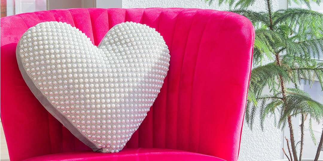Bright&amp;&#x23;x20&#x3B;pink&amp;&#x23;x20&#x3B;velvet&amp;&#x23;x20&#x3B;chair&amp;&#x23;x20&#x3B;holding&amp;&#x23;x20&#x3B;large&amp;&#x23;x20&#x3B;heart-shaped&amp;&#x23;x20&#x3B;throw&amp;&#x23;x20&#x3B;pillow&amp;&#x23;x20&#x3B;covered&amp;&#x23;x20&#x3B;in&amp;&#x23;x20&#x3B;white&amp;&#x23;x20&#x3B;pearls&amp;&#x23;x20&#x3B;or&amp;&#x23;x20&#x3B;rhinestones,&amp;&#x23;x20&#x3B;with&amp;&#x23;x20&#x3B;green&amp;&#x23;x20&#x3B;foliage&amp;&#x23;x20&#x3B;plant&amp;&#x23;x20&#x3B;visible&amp;&#x23;x20&#x3B;beside&amp;&#x23;x20&#x3B;the&amp;&#x23;x20&#x3B;chair.