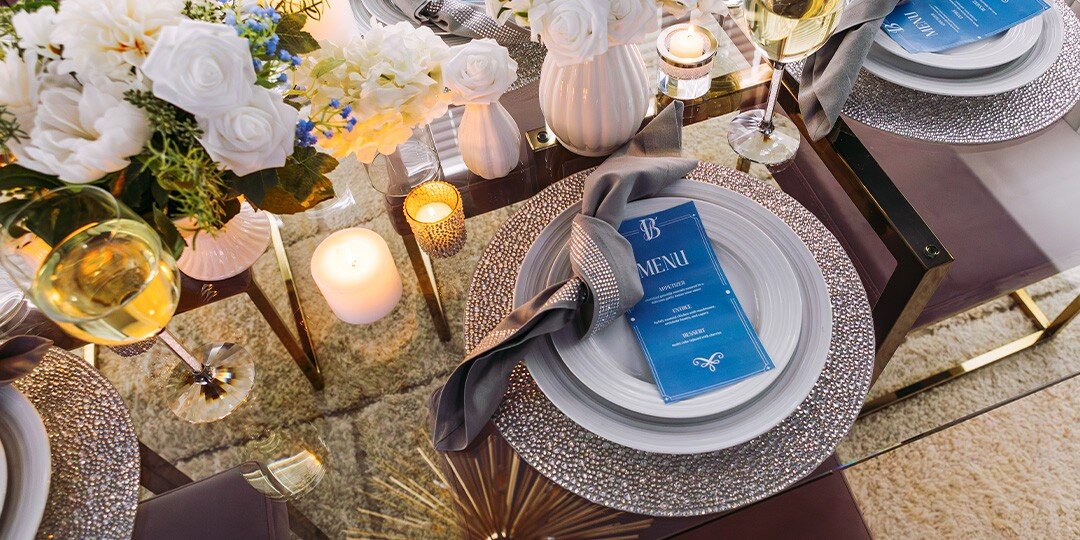 Elegant&amp;&#x23;x20&#x3B;overhead&amp;&#x23;x20&#x3B;view&amp;&#x23;x20&#x3B;of&amp;&#x23;x20&#x3B;formal&amp;&#x23;x20&#x3B;table&amp;&#x23;x20&#x3B;setting&amp;&#x23;x20&#x3B;featuring&amp;&#x23;x20&#x3B;sparkly&amp;&#x23;x20&#x3B;silver&amp;&#x23;x20&#x3B;rhinestone&amp;&#x23;x20&#x3B;charger&amp;&#x23;x20&#x3B;plates,&amp;&#x23;x20&#x3B;white&amp;&#x23;x20&#x3B;dinner&amp;&#x23;x20&#x3B;plates,&amp;&#x23;x20&#x3B;dark&amp;&#x23;x20&#x3B;napkins,&amp;&#x23;x20&#x3B;blue&amp;&#x23;x20&#x3B;menu&amp;&#x23;x20&#x3B;cards,&amp;&#x23;x20&#x3B;lit&amp;&#x23;x20&#x3B;candles,&amp;&#x23;x20&#x3B;white&amp;&#x23;x20&#x3B;rose&amp;&#x23;x20&#x3B;arrangements,&amp;&#x23;x20&#x3B;and&amp;&#x23;x20&#x3B;wine&amp;&#x23;x20&#x3B;glasses&amp;&#x23;x20&#x3B;on&amp;&#x23;x20&#x3B;reflective&amp;&#x23;x20&#x3B;surface.