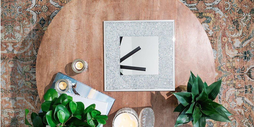 An&amp;&#x23;x20&#x3B;overhead&amp;&#x23;x20&#x3B;view&amp;&#x23;x20&#x3B;of&amp;&#x23;x20&#x3B;a&amp;&#x23;x20&#x3B;round&amp;&#x23;x20&#x3B;wooden&amp;&#x23;x20&#x3B;coffee&amp;&#x23;x20&#x3B;table&amp;&#x23;x20&#x3B;styled&amp;&#x23;x20&#x3B;with&amp;&#x23;x20&#x3B;decorative&amp;&#x23;x20&#x3B;accessories,&amp;&#x23;x20&#x3B;positioned&amp;&#x23;x20&#x3B;on&amp;&#x23;x20&#x3B;a&amp;&#x23;x20&#x3B;vintage&amp;&#x23;x20&#x3B;patterned&amp;&#x23;x20&#x3B;area&amp;&#x23;x20&#x3B;rug&amp;&#x23;x20&#x3B;in&amp;&#x23;x20&#x3B;earth&amp;&#x23;x20&#x3B;tones.&amp;&#x23;x20&#x3B;The&amp;&#x23;x20&#x3B;table&amp;&#x23;x20&#x3B;features&amp;&#x23;x20&#x3B;a&amp;&#x23;x20&#x3B;square&amp;&#x23;x20&#x3B;mirrored&amp;&#x23;x20&#x3B;tray&amp;&#x23;x20&#x3B;bordered&amp;&#x23;x20&#x3B;in&amp;&#x23;x20&#x3B;tiny&amp;&#x23;x20&#x3B;silver&amp;&#x23;x20&#x3B;rhinestones&amp;&#x23;x20&#x3B;under&amp;&#x23;x20&#x3B;the&amp;&#x23;x20&#x3B;glass,&amp;&#x23;x20&#x3B;flanked&amp;&#x23;x20&#x3B;by&amp;&#x23;x20&#x3B;green&amp;&#x23;x20&#x3B;potted&amp;&#x23;x20&#x3B;plants,&amp;&#x23;x20&#x3B;small&amp;&#x23;x20&#x3B;lit&amp;&#x23;x20&#x3B;candles&amp;&#x23;x20&#x3B;in&amp;&#x23;x20&#x3B;glass&amp;&#x23;x20&#x3B;holders,&amp;&#x23;x20&#x3B;and&amp;&#x23;x20&#x3B;a&amp;&#x23;x20&#x3B;book.&amp;&#x23;x20&#x3B;The&amp;&#x23;x20&#x3B;composition&amp;&#x23;x20&#x3B;creates&amp;&#x23;x20&#x3B;a&amp;&#x23;x20&#x3B;curated,&amp;&#x23;x20&#x3B;modern&amp;&#x23;x20&#x3B;living&amp;&#x23;x20&#x3B;space&amp;&#x23;x20&#x3B;vignette.