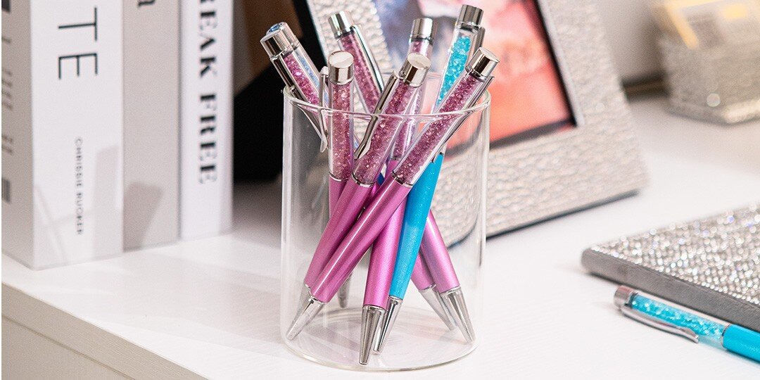 Clear&amp;&#x23;x20&#x3B;glass&amp;&#x23;x20&#x3B;pen&amp;&#x23;x20&#x3B;holder&amp;&#x23;x20&#x3B;filled&amp;&#x23;x20&#x3B;with&amp;&#x23;x20&#x3B;colorful&amp;&#x23;x20&#x3B;pens&amp;&#x23;x20&#x3B;featuring&amp;&#x23;x20&#x3B;rhinestone-topped&amp;&#x23;x20&#x3B;caps&amp;&#x23;x20&#x3B;in&amp;&#x23;x20&#x3B;pink,&amp;&#x23;x20&#x3B;blue,&amp;&#x23;x20&#x3B;and&amp;&#x23;x20&#x3B;other&amp;&#x23;x20&#x3B;colors,&amp;&#x23;x20&#x3B;positioned&amp;&#x23;x20&#x3B;on&amp;&#x23;x20&#x3B;white&amp;&#x23;x20&#x3B;desk&amp;&#x23;x20&#x3B;next&amp;&#x23;x20&#x3B;to&amp;&#x23;x20&#x3B;books&amp;&#x23;x20&#x3B;and&amp;&#x23;x20&#x3B;sparkly&amp;&#x23;x20&#x3B;accessories.