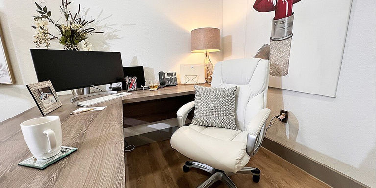 Una oficina en casa moderna con un escritorio de madera clara con dos monitores negros, una silla de oficina ergonómica blanca con un cojín de diamantes de imitación plateados, y una lámpara de mesa cálida con una pantalla beige. El espacio de trabajo incluye suministros de oficina, una disposición de plantas decorativas y obras de arte contemporáneas en paredes blancas. Los pisos de madera oscura completan la estética limpia y profesional.