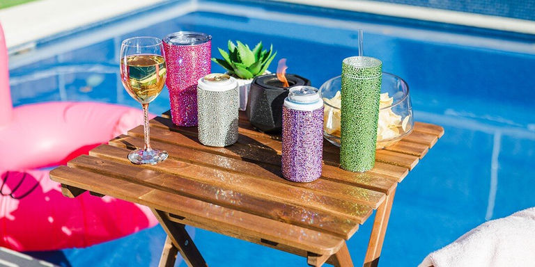 Poolside wooden table displaying collection of rhinestone drink accessories including pink, silver, green, and purple sparkly can coolers and tumblers, wine glass, and small succulent plant, with blue swimming pool and pink float visible in background.