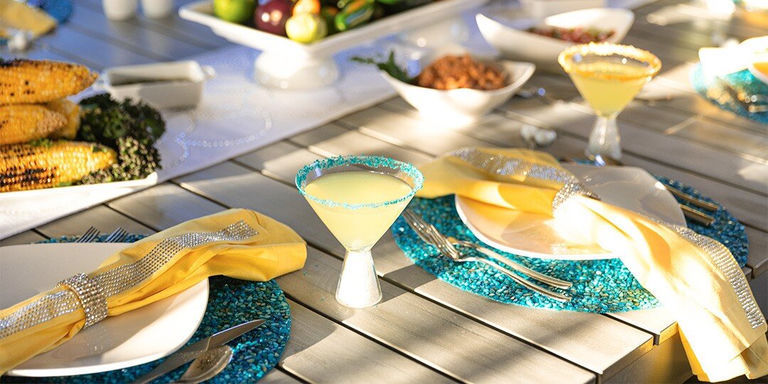 Vibrant outdoor dining table with bright pink tablecloth, place settings featuring sparkly round placemats, white dinnerware, yellow napkins, and colorful floral centerpiece, set against brick wall garden backdrop.