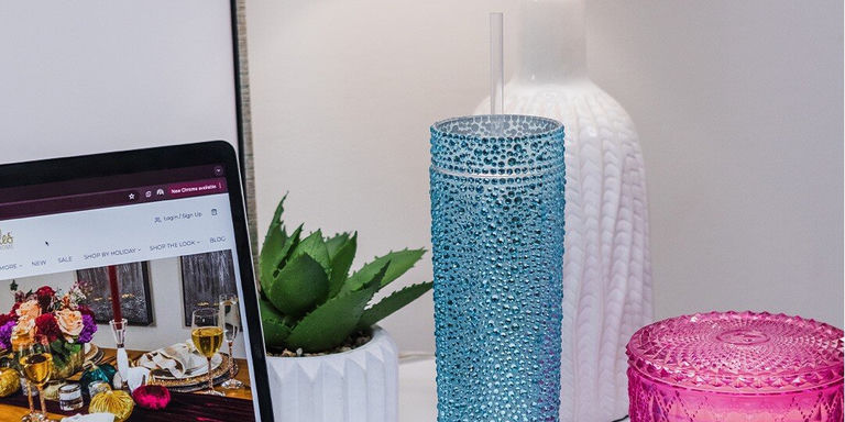 Desk workspace featuring laptop displaying website, blue rhinestone tumbler with straw, small succulent plant in white pot, and pink rhinestone container, with white textured vase in background.