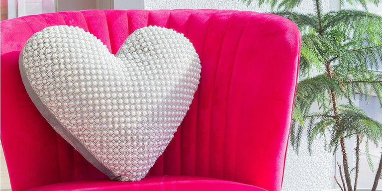 Bright pink velvet chair holding large heart-shaped throw pillow covered in white pearls or rhinestones, with green foliage plant visible beside the chair.
