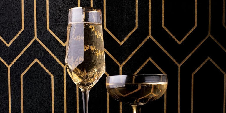 Champagne flute and coupe glass in front of black and gold Art Deco wallpaper pattern