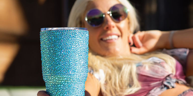 Femme se prélassant en plein air tenant un gobelet bleu en strass, portant des lunettes de soleil glamour