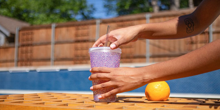 Lavender rhinestone tumbler held by hands at poolside with orange