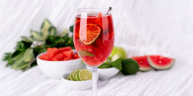 red watermelon margarita garnished with citrus and mint surrounded by fresh fruit on white background