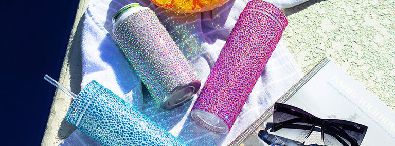 Colorful rhinestone-wrapped tumblers in blue, pink, and turquoise lying on white towel beside pool with sunglasses