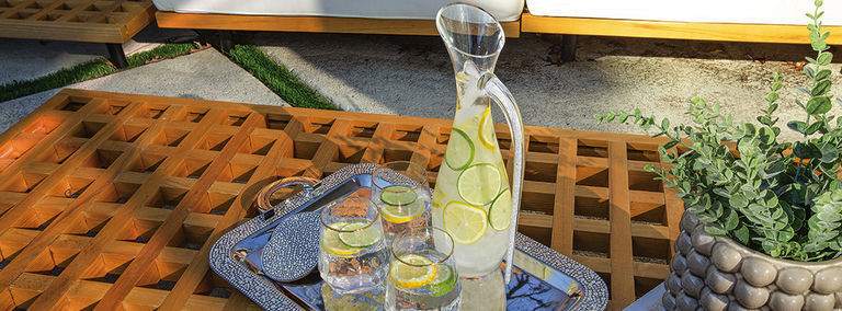 Outdoor teak table with rhinestone-wrapped serving tray holding citrus water pitcher and glasses with lime slices beside pool