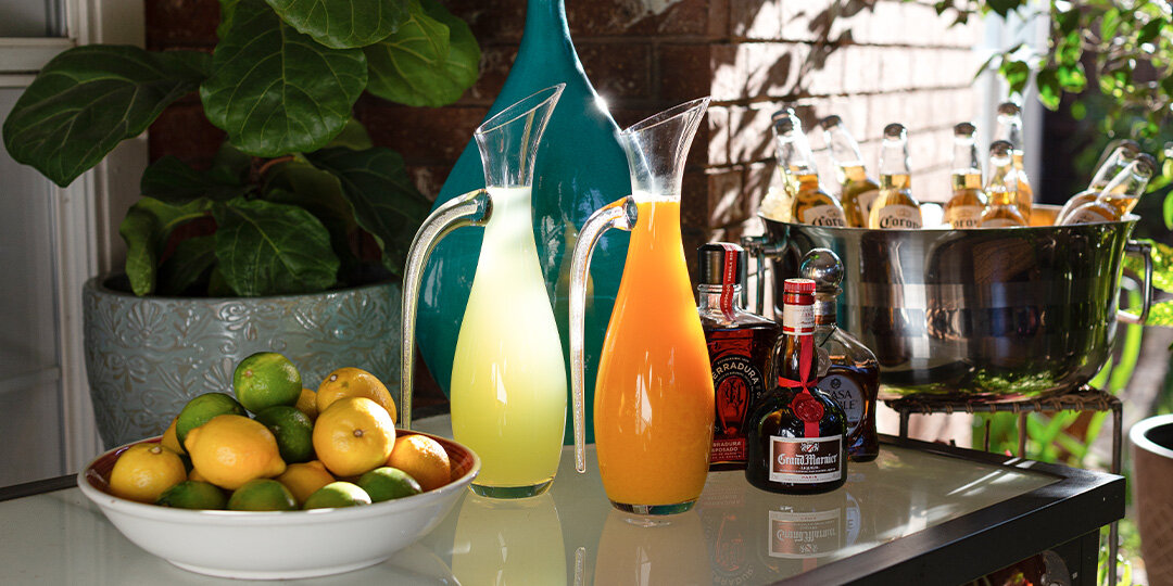 Outdoor&amp;&#x23;x20&#x3B;entertaining&amp;&#x23;x20&#x3B;setup&amp;&#x23;x20&#x3B;on&amp;&#x23;x20&#x3B;a&amp;&#x23;x20&#x3B;kitchen&amp;&#x23;x20&#x3B;counter&amp;&#x23;x20&#x3B;featuring&amp;&#x23;x20&#x3B;two&amp;&#x23;x20&#x3B;glass&amp;&#x23;x20&#x3B;pitchers&amp;&#x23;x20&#x3B;with&amp;&#x23;x20&#x3B;yellow&amp;&#x23;x20&#x3B;and&amp;&#x23;x20&#x3B;orange&amp;&#x23;x20&#x3B;beverages,&amp;&#x23;x20&#x3B;white&amp;&#x23;x20&#x3B;bowl&amp;&#x23;x20&#x3B;filled&amp;&#x23;x20&#x3B;with&amp;&#x23;x20&#x3B;mixed&amp;&#x23;x20&#x3B;citrus&amp;&#x23;x20&#x3B;fruits&amp;&#x23;x20&#x3B;&amp;&#x23;x28&#x3B;limes,&amp;&#x23;x20&#x3B;lemons,&amp;&#x23;x20&#x3B;oranges&amp;&#x23;x29&#x3B;,&amp;&#x23;x20&#x3B;dark&amp;&#x23;x20&#x3B;liquor&amp;&#x23;x20&#x3B;bottles,&amp;&#x23;x20&#x3B;ice&amp;&#x23;x20&#x3B;bucket&amp;&#x23;x20&#x3B;with&amp;&#x23;x20&#x3B;beer&amp;&#x23;x20&#x3B;bottles,&amp;&#x23;x20&#x3B;and&amp;&#x23;x20&#x3B;large&amp;&#x23;x20&#x3B;teal&amp;&#x23;x20&#x3B;decorative&amp;&#x23;x20&#x3B;vase.&amp;&#x23;x20&#x3B;Large&amp;&#x23;x20&#x3B;green&amp;&#x23;x20&#x3B;plant&amp;&#x23;x20&#x3B;and&amp;&#x23;x20&#x3B;patio&amp;&#x23;x20&#x3B;setting&amp;&#x23;x20&#x3B;visible&amp;&#x23;x20&#x3B;in&amp;&#x23;x20&#x3B;background.