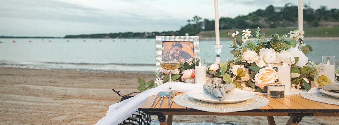 Romantic&amp;&#x23;x20&#x3B;beachside&amp;&#x23;x20&#x3B;table&amp;&#x23;x20&#x3B;setting&amp;&#x23;x20&#x3B;on&amp;&#x23;x20&#x3B;wooden&amp;&#x23;x20&#x3B;table&amp;&#x23;x20&#x3B;featuring&amp;&#x23;x20&#x3B;sparkly&amp;&#x23;x20&#x3B;rhinestone&amp;&#x23;x20&#x3B;charger&amp;&#x23;x20&#x3B;plates,&amp;&#x23;x20&#x3B;white&amp;&#x23;x20&#x3B;candles,&amp;&#x23;x20&#x3B;cream&amp;&#x23;x20&#x3B;and&amp;&#x23;x20&#x3B;peach&amp;&#x23;x20&#x3B;floral&amp;&#x23;x20&#x3B;centerpiece,&amp;&#x23;x20&#x3B;wine&amp;&#x23;x20&#x3B;glasses,&amp;&#x23;x20&#x3B;and&amp;&#x23;x20&#x3B;rhinestone&amp;&#x23;x20&#x3B;picture&amp;&#x23;x20&#x3B;frame&amp;&#x23;x20&#x3B;displaying&amp;&#x23;x20&#x3B;a&amp;&#x23;x20&#x3B;couple
